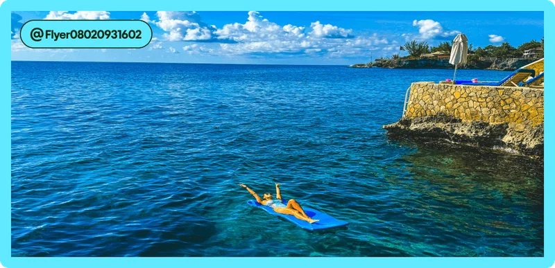Image Description: A person laying out on a raft in the middle of the ocean, throwing their hands in the air. A cobblestoned wall with lounge chairs can be seen just to their right.