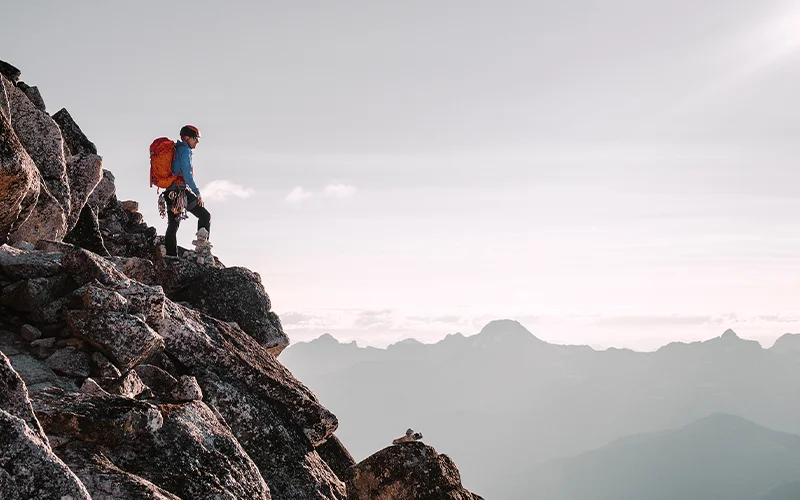 A mountain climber—perched on Bugaboo Spire in Bugaboo National Park—looks out at the sun with the Purcell Mountain Range in the background.