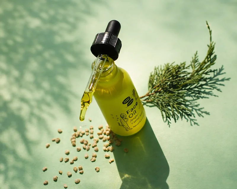 A green bottle of face oil on a green background stands with a dropper full of oil balanced on the top. A sprig of juniper and brown seeds are strewn around the bottle.