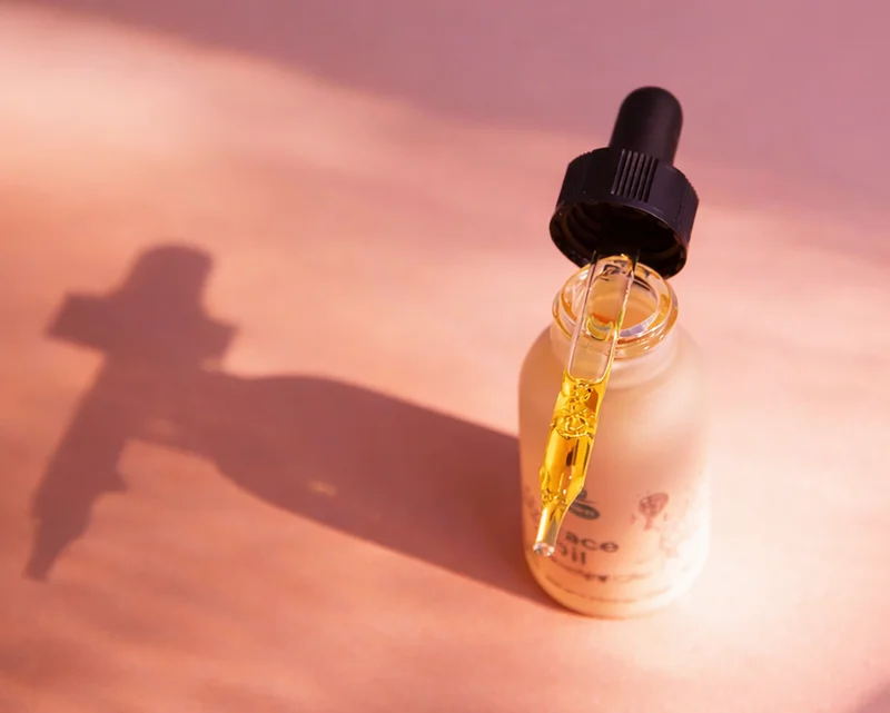 A pink bottle of face oil on a pink background stands with a dropper full of oil balanced on the top