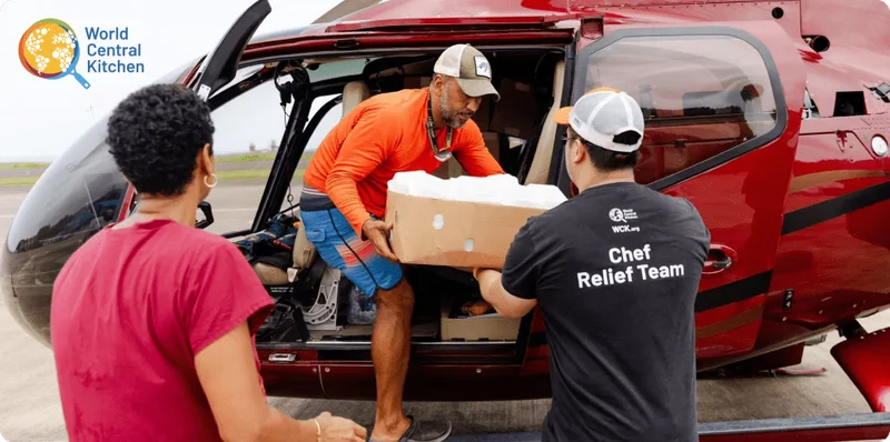 Image Description: Man unloads a box off a helicopter, handing it to another man wearing a World Central Kitchen t-shirt that says ‘Chef Relief Team’ on the back. A woman stands to the side waiting to assist.