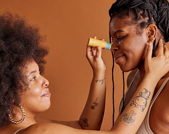 A Black woman with an afro applies repair balm to the nose of another Black woman, sweetly. smiles are on both faces