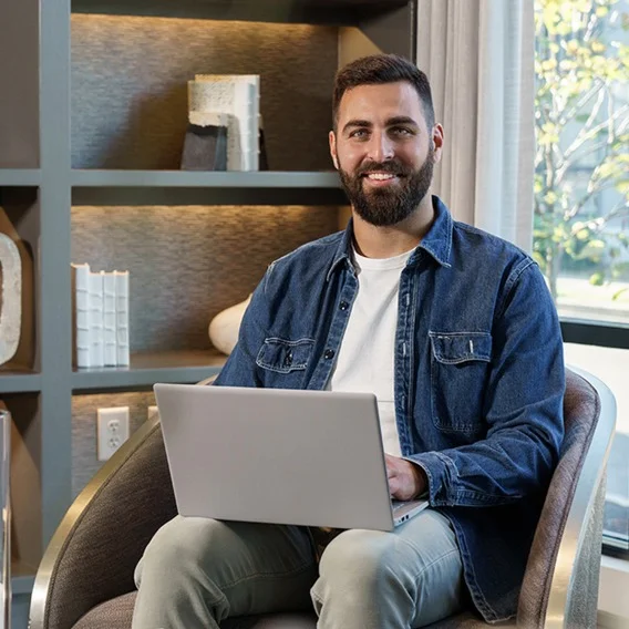 A man with tan skin and a beard, dressed in a blue button down shirt, sits in a chair in front of a bookshelf holding a laptop computer.