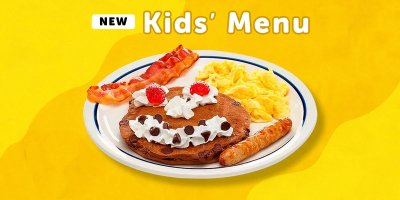 The Happy Face Combo from IHOP’s Kids’ Menu, featuring scrambled eggs, a bacon strip, a sausage link, and a pancake with a face: Maraschino cherry eyes and a whipped topping nose and mouth with some chocolate chips for teeth.