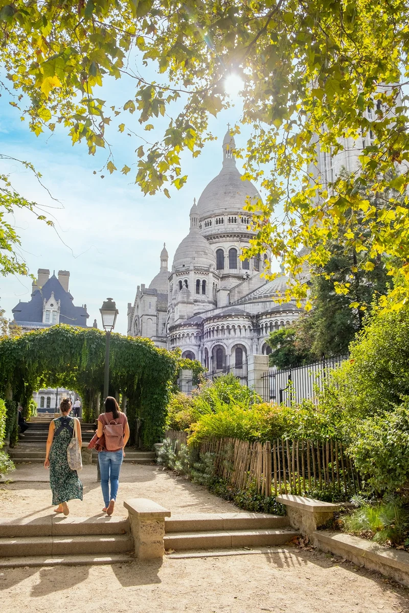 Two women strolling through a Parisian garden with iconic domed buildings in the back