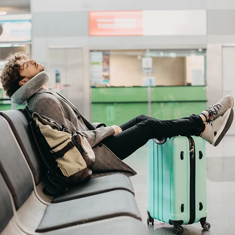 Young businessman sleeps alone in the waiting room of a station. He sits with his legs on suitcase and a pillow around his neck.