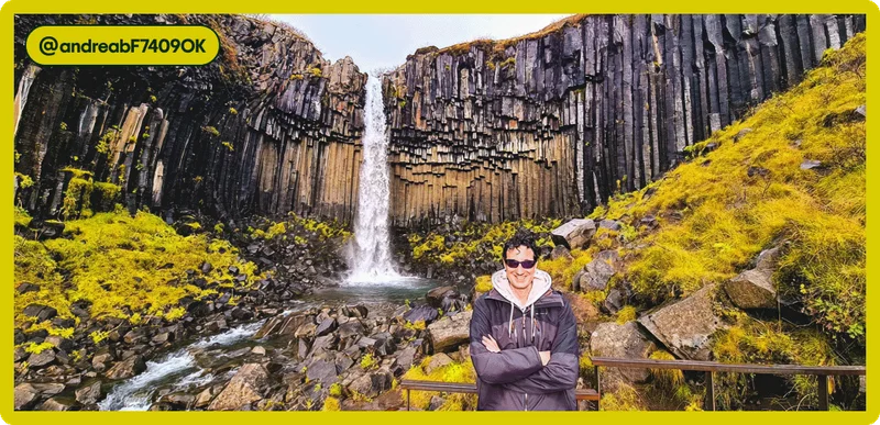 Image Description: Person poses for a photo in front of a large waterfall with abnormal rock formations and mossy green rocks below. Tag attribution in the corner for user @andreabF7409OK.