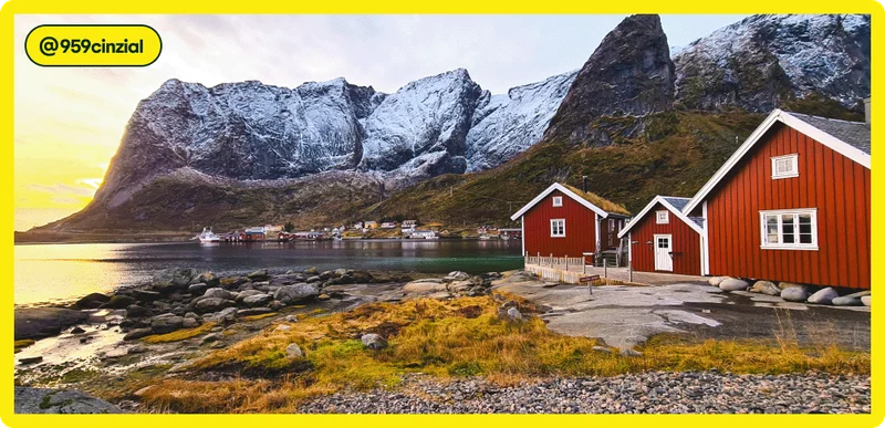 Image Description: Scenic landscape photo showing a couple of red houses next to the water with a rocky coastline and snow-covered mountains in the background. The sun sets just beyond the mountains. Tag attribution in the corner for user @959cinzial.