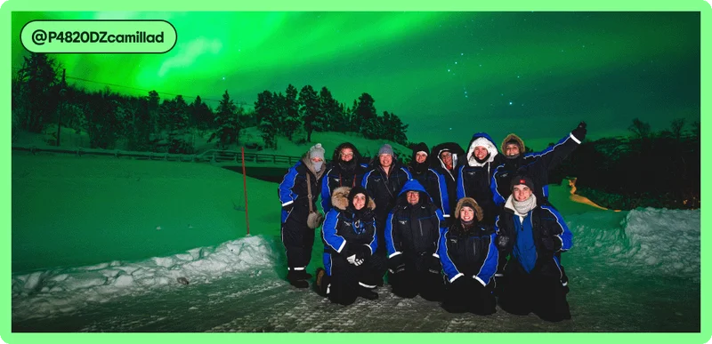 Image Description: Group tour in matching snowsuits posing for a photo at night with the green glow of the northern lights behind them. Tag attribution in the corner for user @P4820DZcamillad.