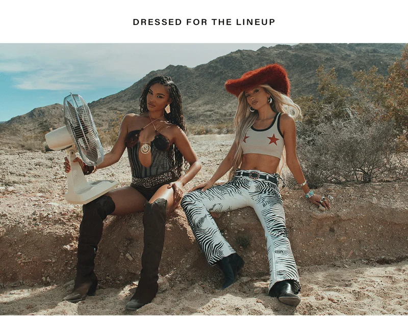Two women in bold festival fashion, desert setting. One in dark corset, other in red fuzzy hat and zebra pants. Shop concert outfits.