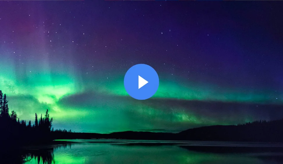 The Northern Lights dance across the night sky, painting it with vibrant green and purple hues. A calm lake reflects the colorful aurora and the silhouettes of evergreen trees beneath a starry sky.
