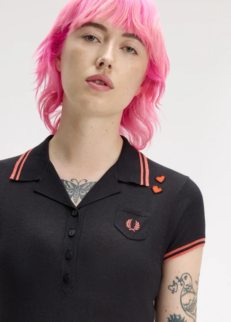 Fred Perry x The Amy Winehouse Foundation: female model with choppy neck-length pink hair looks directly into the camera and wears a black polo with a revere collar and coral pink tipping.