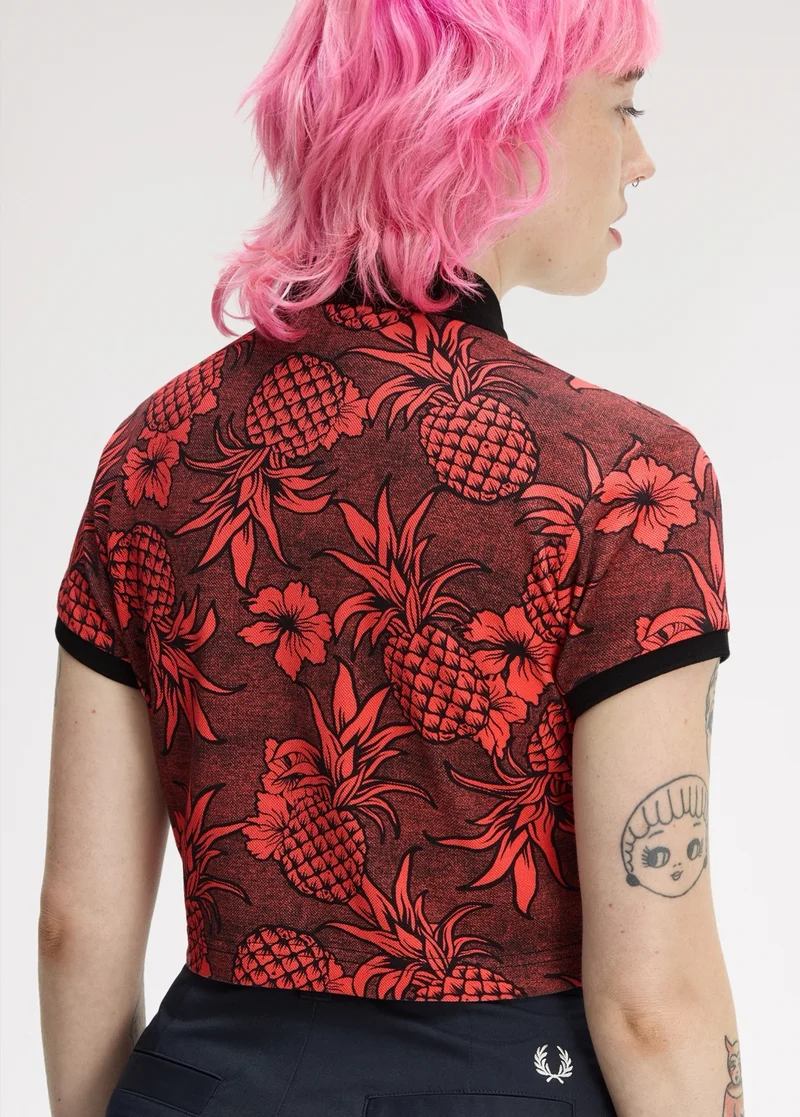 Fred Perry x The Amy Winehouse Foundation: female model with choppy neck-length pink hair looks away from the camera and wear a cropped polo shirt. The polo is has a pattern, reminiscent of traditional tattoo style, in coral pink and black featuring hibiscus flows and pineapples.
