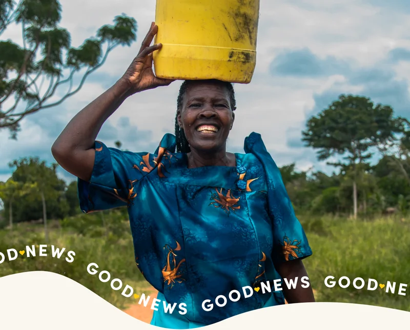 A woman carries a yellow jerry can of water on her head while smiling.