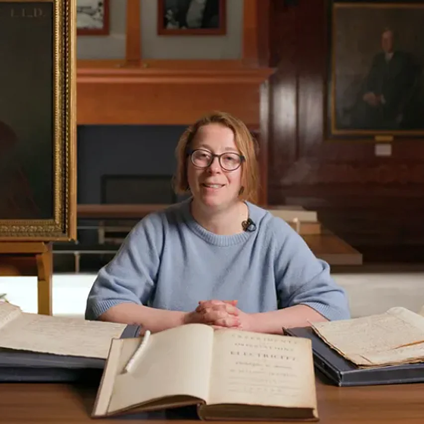 A woman in glasses smiles behind a desk featuring open antique books. The central text reads 'Experiments and Observations on Electricity.' Framed portraits hang in the wood-paneled room behind her.