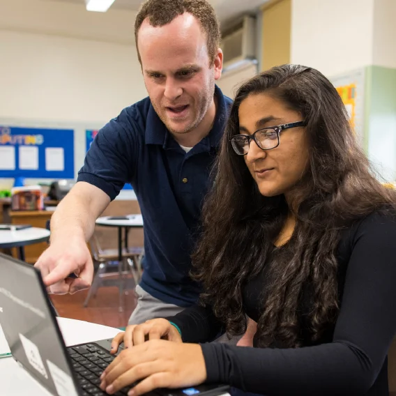 A man is leaning over and pointing at a laptop screen while a woman with glasses and long dark hair types on the keyboard