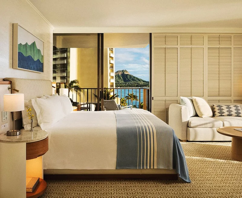 Image Description: A white bed with a blue throw blanket in an airy hotel room with a balcony overlooking the beach and an island in the distance.
