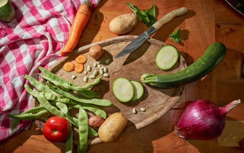 An array of vegetables like carrots, snap peas, tomatoes, and zucchini are displayed on a wooden cutting board.