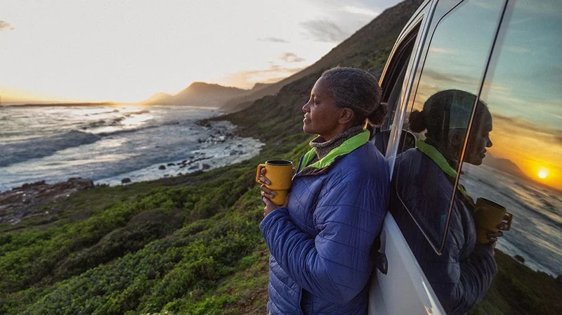 Image description: Woman standing next to car with a cup of coffee watching the sunset over the ocean