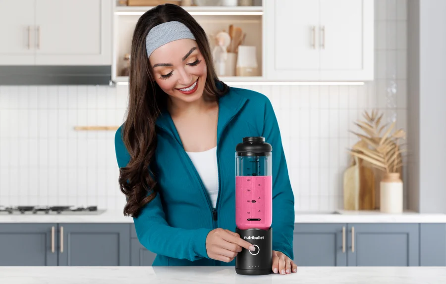 A woman in sports wear using a nutribullet blender with pink liquid in a kitchen setting.