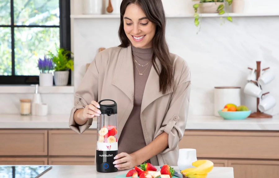 A woman using a portable nutribullet blender in a bright kitchen with fruits on the counter.