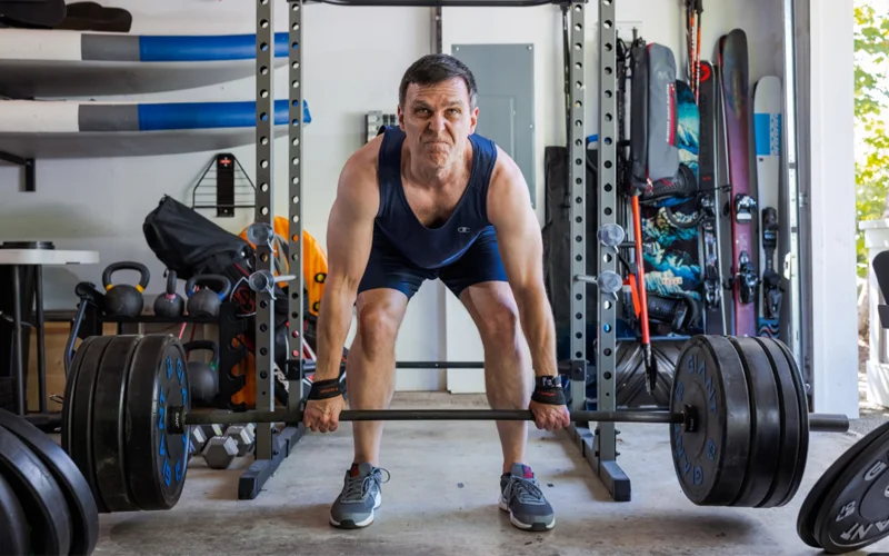 An older man lifts barbell weights in a garage.