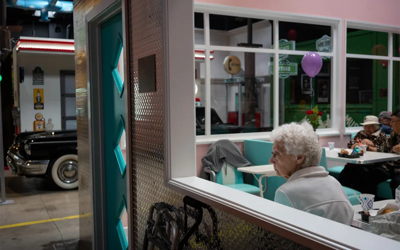 A octogenarian peers out of a 1950s-style diner, one of many nostalgic settings at Glenner Town Square, a dementia day care center in Chula Vista, California.