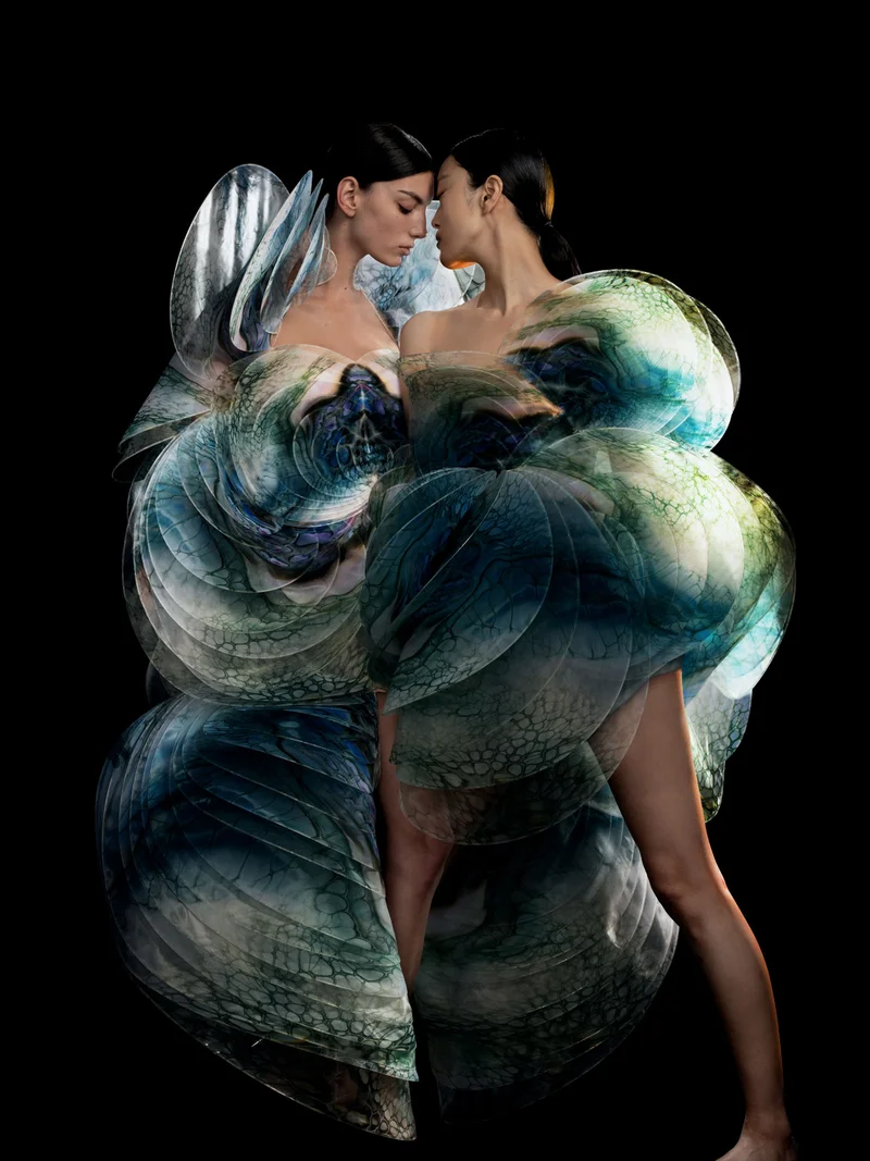 Two models facing each other, wearing layered sculptural garments