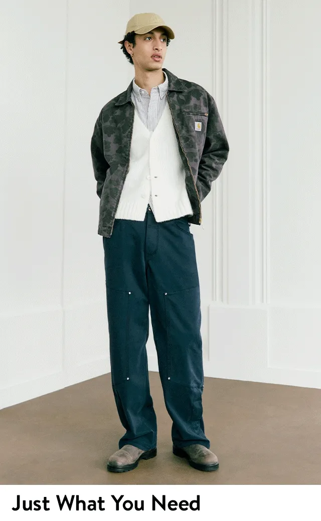 A man wearing a workwear jacket over a button-shirt and knit vest with utility pants and boots.