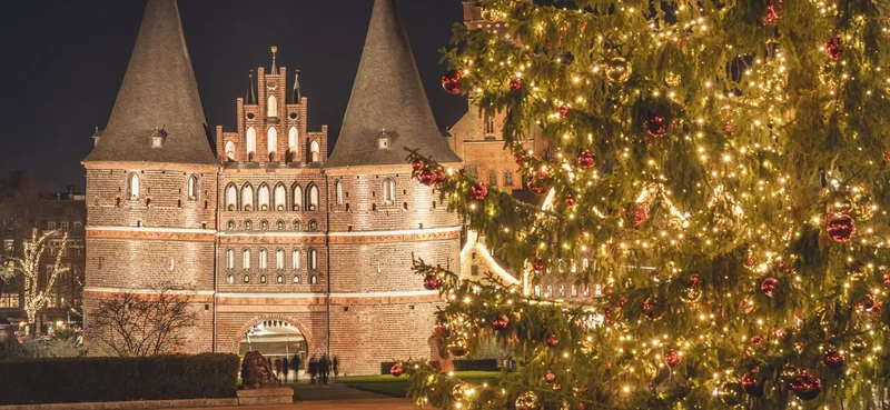 Illuminated Holstentor gate at night in Lubeck, Germany, adorned with a glowing Christmas tree in the foreground.