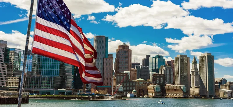 Boston skyline featuring iconic skyscrapers and landmarks, with the United States national flag in the foreground.