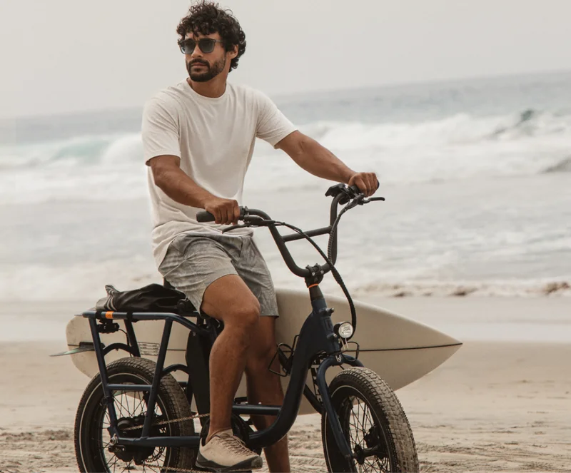 Man riding a bike on the beach with a surfboard, wearing casual coastal outfit under headline “The all-day explorer.”