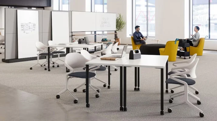 Two people sitting in a brightly lit, open floorplan community setting featuring Herman Miller Fuld Nesting chairs, yellow NaughtOne Pippin chairs, and Herman Miller OE1 Agile Walls.