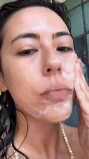 A person with brown hair and tan skin applies cleanser, spray toner and face oil to their face while outside among the trees.