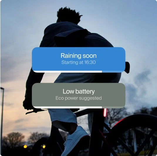 Silhouette of a rider on a Cowboy bike at dusk with the app displaying safety alerts: “Raining soon” and “Low battery - Eco power suggested.”