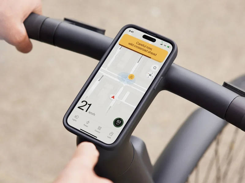Close-up of a Cowboy bike’s handlebars with a smartphone mounted on the stem, displaying the app interface with navigation and a warning message reading “Careful now, wild crossroad ahead.” The screen also shows a speed of 21 km/h.