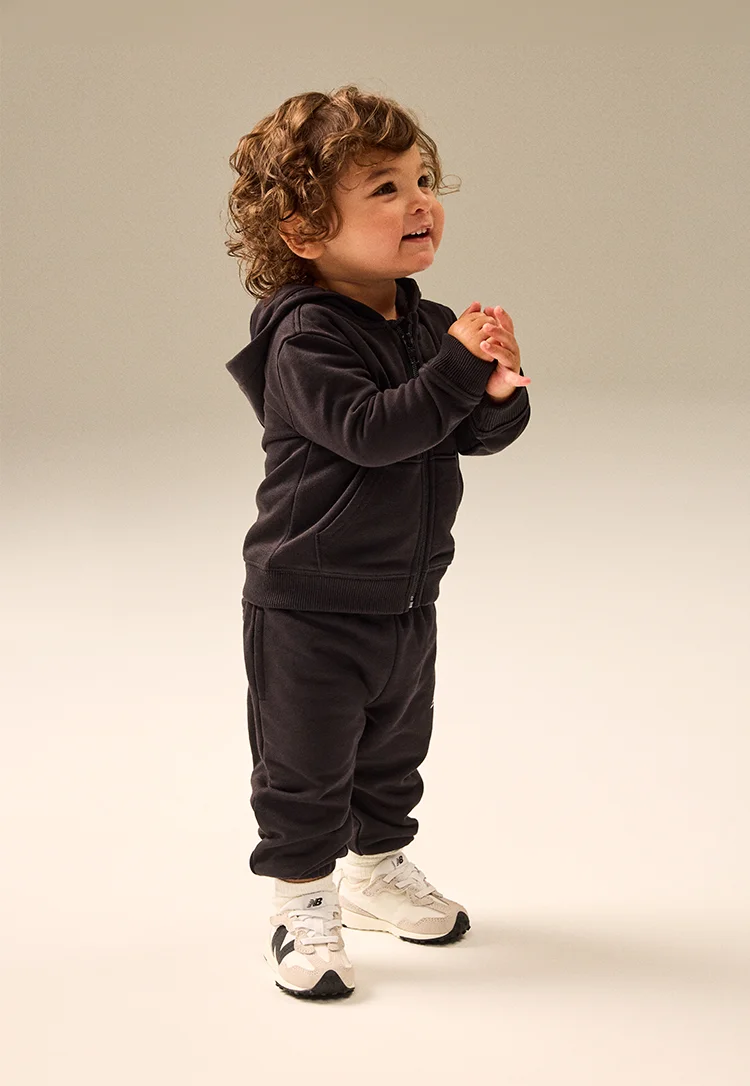 A toddler with brown curls, delighting in their all black hoodie and sweatpants, and tiny white and black New Balance shoes
