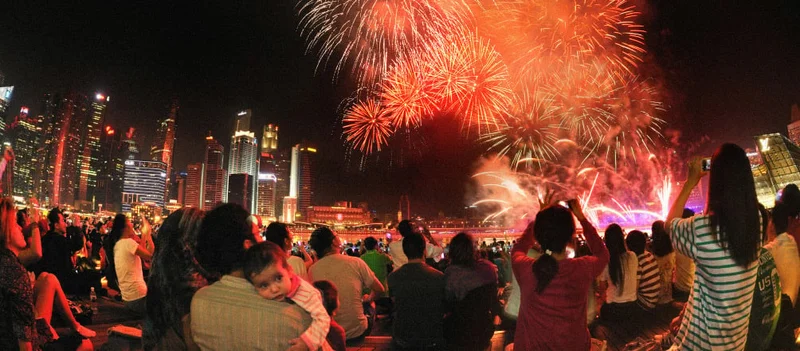 Image Description: A crowd of travelers watching and taking photos of fireworks at Marina Bay from an outdoor plaza