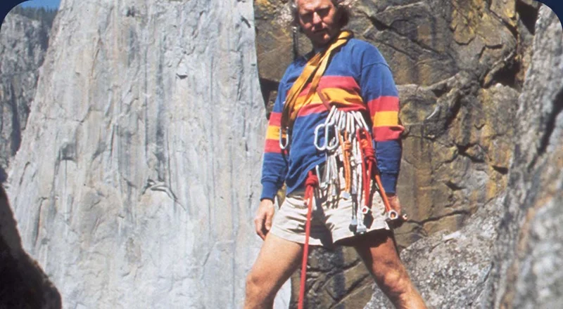 A person wearing a rugby shirt, shorts and climbing gear strikes a dramatic pose in front of a granite wall.