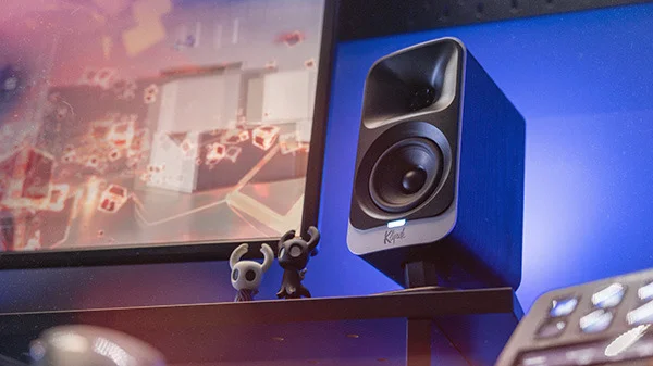 The Klipsch ProMedia Lumina sitting on the raised bar of a gaming desk next to two small video game figurines. The lights from the speaker cast a cool blue glow on the back wall. .