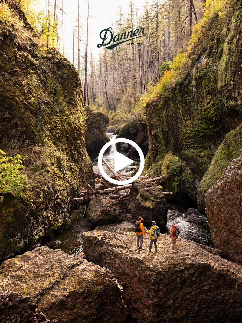 Three hikers stand on a rocky outcrop in a mossy canyon with a river. A play button is centered on the image, with the Danner logo at the top.