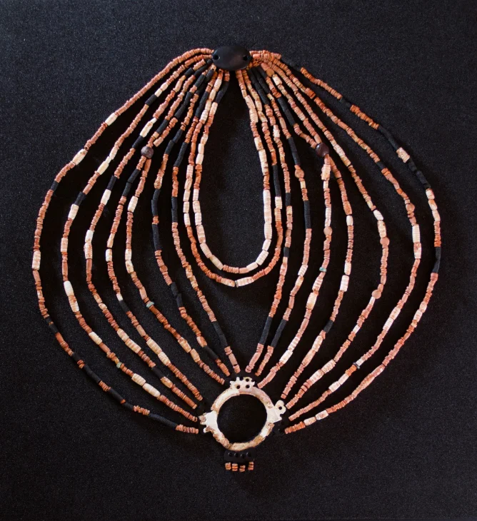 This Neolithic-era necklace is fashioned from sea shells, turquoise, amber, and more, with an engraved mother-of-pearl ring and a hematite pendant. Black foam tubes are used to indicate the presence of beads too poorly preserved to be displayed.