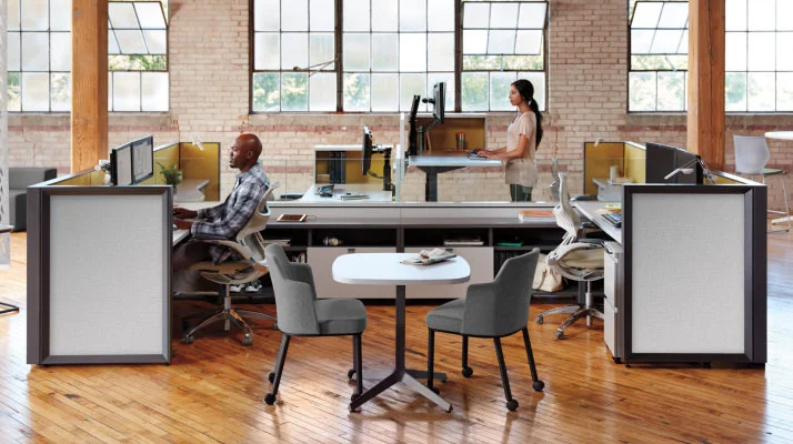 A group of workstations in an office with one man in the foreground seated and working at a desk and facing left and a woman in the background standing and working at a desk also facing left.