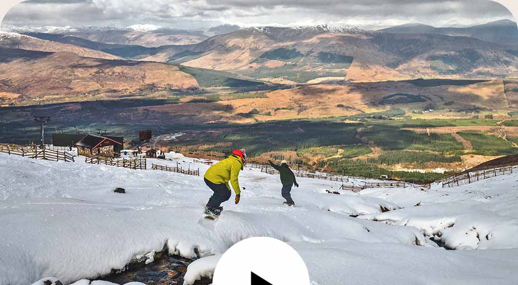 Video play button. Two snowboarders ride down a snowy hill with a stream running through it.