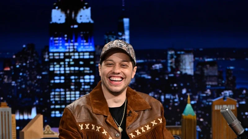 Image may contain: Pete Davidson, Clothing, Pants, Coat, Jacket, Person, Sitting, Footwear, Shoe, Hat, and Teen
