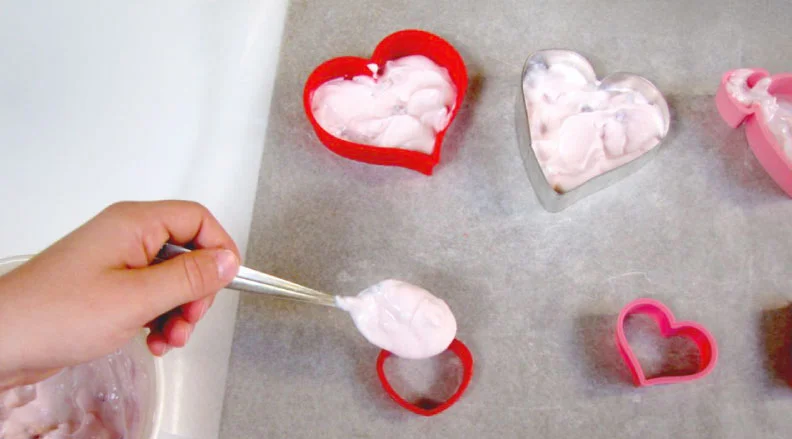 A hand placing a yogurt-based heart-shaped treat on parchment paper, surrounded by other pink and white yogurt hearts.