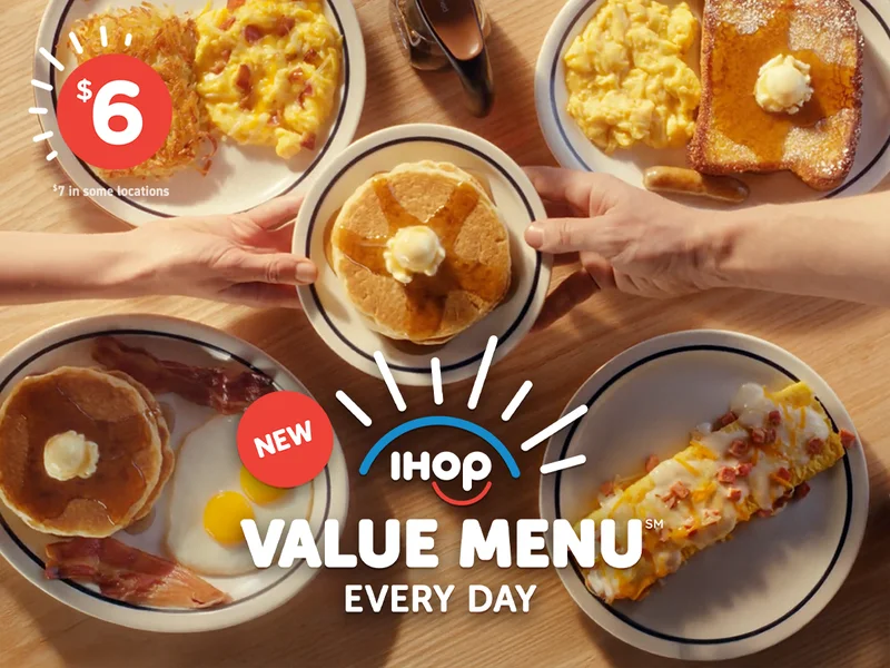 A wooden table has four Value Menu items on it, including the Breakfast Faves Combo and Ham & Cheese Omelette. Two different hands reach across from the left and right of the table and hold a plate of buttermilk pancakes topped with syrup and butter.