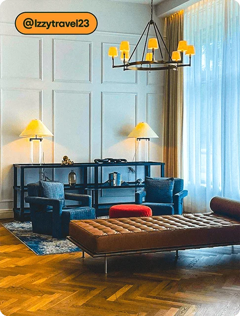 Image description: A sitting room at a hotel with white walls and a floor-to-ceiling window with sheer drapes. There’s a bookshelf with two lamps, two cozy chairs with a red ottoman between them, and brown leather chaise. A modern lighting fixture hangs above it all.