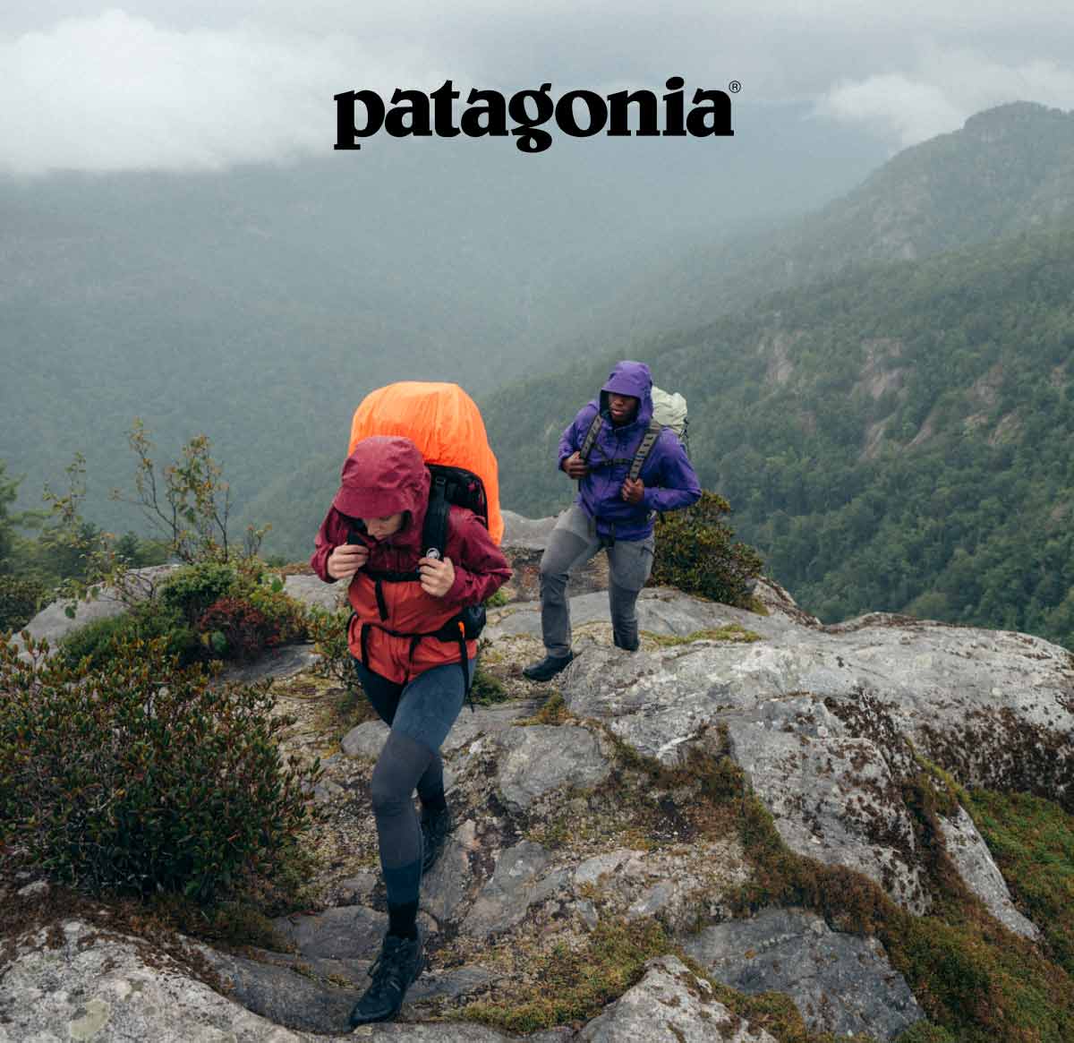 Patagonia logo. Two people wearing large backpacks and rain gear hike over a rocky ridge with mountains and cloudy skies in the background.
