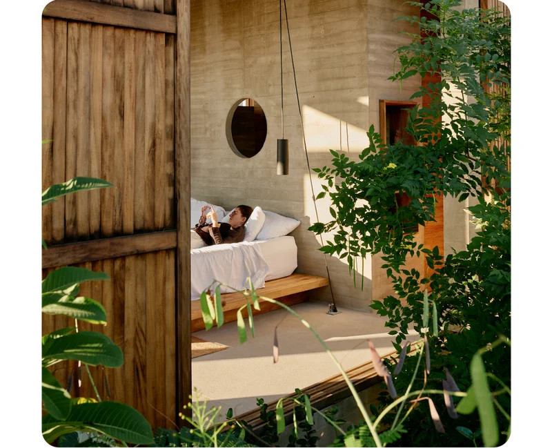 Two large, wood-slatted windows open to reveal a modern, concrete interior. A person lying on a platform bed with white linens watches their phone as daylight spills in from an unseen source. Greenery surrounds the space outside.